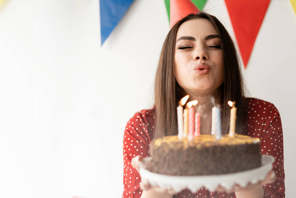 Young beautiful stylish woman holds a cake in her hands and blows out candles on it. Concept of making a wish in honor of the holiday. Evening party at home - Фото, зображення