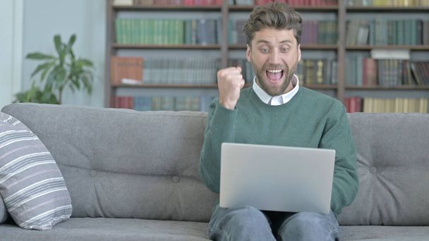 Úspěšný muž pracovat na přenosném počítači a slavit během sezení na pohovce - Fotografie, Obrázek