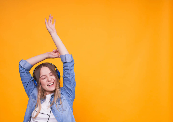 Portrait of a girl in casual clothes who is happy to listen to music on headphones, rejoicing, dancing, having fun with raised hands on a yellow background. Female student with closed eyes,copy space. - Foto, Bild