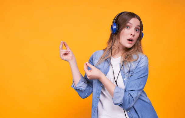 Portrait of a Caucasian girl who enjoys listening to music on headphones, is surprised, claps her fingers, rejoicing, dancing, having fun on a yellow background. Female student copy space. - Fotoğraf, Görsel