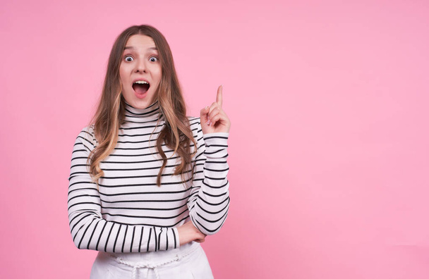 Emotional young surprised fair-haired lady looks with saggy eyes, feels something impressed, points with her index finger, dressed in a casual striped sweater isolated on pink background, copy space. - Valokuva, kuva