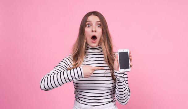 Portret van een mooi jong blank meisje met verrassing en shock toont de wijsvinger van haar linkerhand op SMS aan de telefoon, in een gestreepte basic zwart-wit trui op een roze achtergrond. - Foto, afbeelding