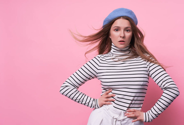Portrait of a cute emotional passionate Caucasian girl, in a stylish fashionable beret hat with a calm face, holding hands on her hips, the wind develops long blond hair, striped sweater copy space. - Фото, изображение