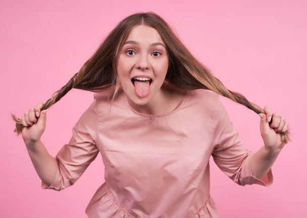 A young Caucasian girl shows her tongue with a funny facial expression, grimaces, holds and stretches her hair with her hands in different directions, fooling around with her hair, pink background. - Fotoğraf, Görsel