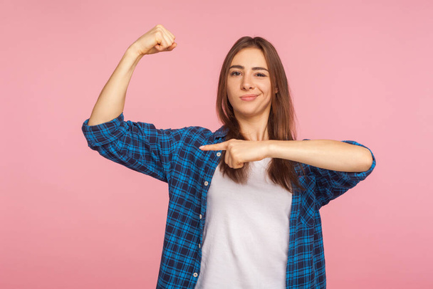 Look, I'm strong! Portrait of haughty successful girl in checkered shirt pointing at biceps and looking with arrogant expression, feeling power, confidence. studio shot isolated on pink background - Valokuva, kuva