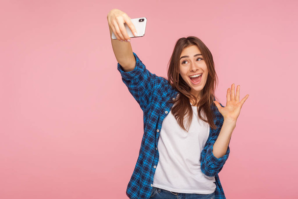 Communication on mobile phone. Portrait of cute cheerful girl in checkered shirt smiling and gesturing hello while having positive talk on video call, online chatting. indoor studio shot, isolated - Photo, Image