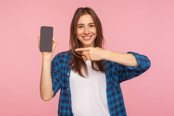 App, mobile device advertising! Happy smiling girl in checkered shirt pointing at cellphone screen, showing display, mock up for commercial text. studio shot isolated on pink background, copy space - Foto, imagen