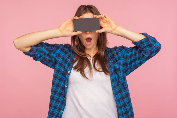 Unbelievable advertisement on mobile device! Amazed girl in checkered shirt covering eyes with smartphone and keeping mouth open in surprise, hiding half face, expressing shock. studio shot, isolated - Photo, image