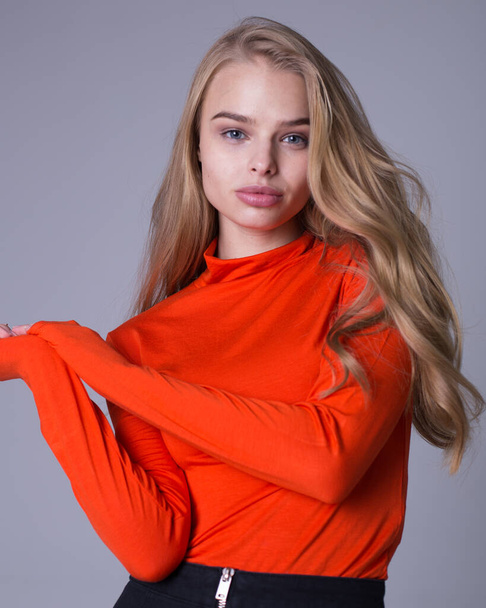 Nice portrait of well-dressed fashionable girl in orange sweatshirt. Lovely lady posing indoors on gray background. Blonde model with serious look. Fashion and lifestyle concept - Zdjęcie, obraz