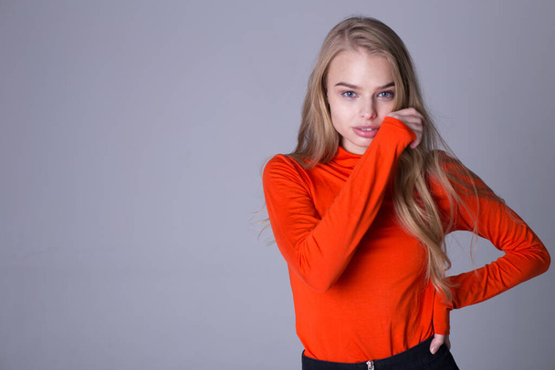 Nice portrait of well-dressed fashionable girl in orange sweatshirt. Lovely lady posing indoors on gray background. Blonde model with serious look. Fashion and lifestyle concept - Fotó, kép