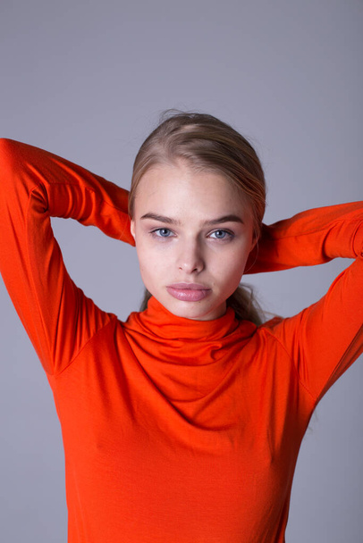 Nice portrait of well-dressed fashionable girl in orange sweatshirt. Lovely lady posing indoors on gray background. Blonde model with serious look. Fashion and lifestyle concept - 写真・画像