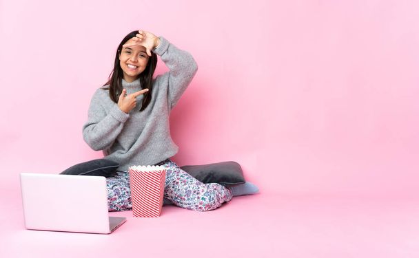 Young mixed race woman eating popcorn while watching a movie on the laptop focusing face. Framing symbol - Photo, Image