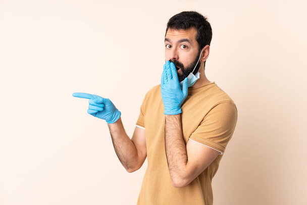 Caucasian man with beard protecting from the coronavirus with a mask and gloves over isolated background with surprise expression while pointing side - Photo, Image