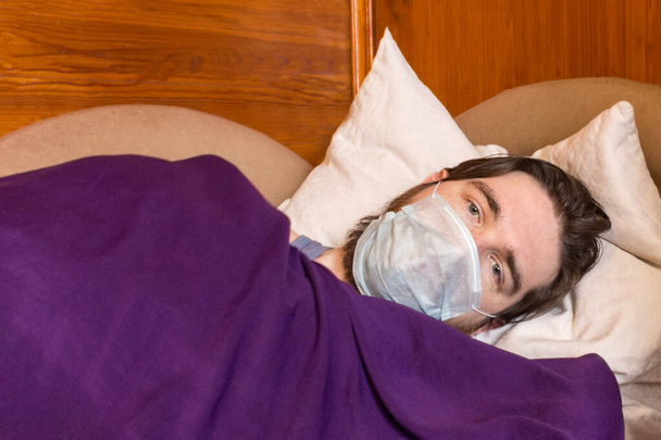 Ένας άντρας με σκούρα μαλλιά και μια μάσκα μιας χρήσης για προστασία από τον ιό, βρίσκεται στο κρεβάτι, απομονώνοντας τον εαυτό του από άλλους ανθρώπους. πυρετός, πονοκέφαλος και ναυτία είναι συμπτώματα - φροντίστε την υγεία σας - Φωτογραφία, εικόνα