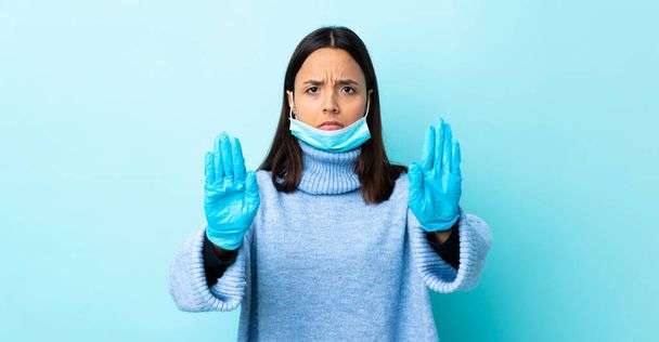 Jovem morena mista mulher protegendo do coronavírus com uma máscara e luvas sobre fundo azul isolado fazendo gesto de parada e decepcionado
 - Foto, Imagem
