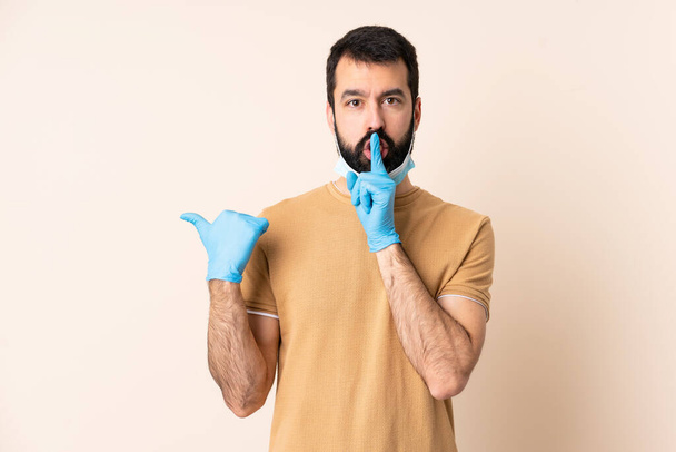 Caucasian man with beard protecting from the coronavirus with a mask and gloves over isolated background pointing to the side and doing silence gesture - Foto, Bild