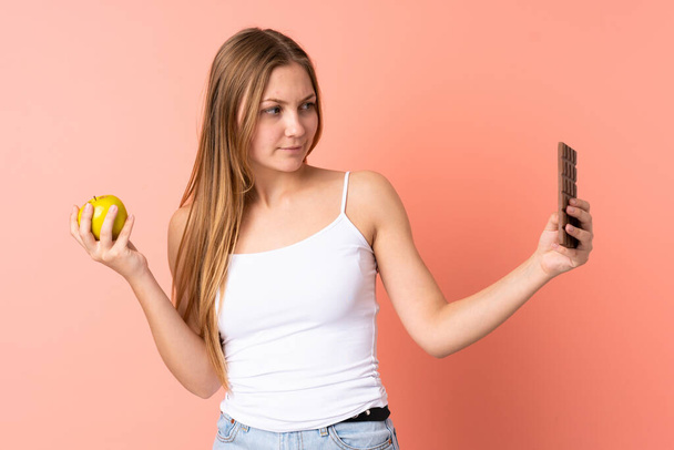 Teenager Ukrainian girl isolated on pink background having doubts while taking a chocolate tablet in one hand and an apple in the other - Foto, Imagen