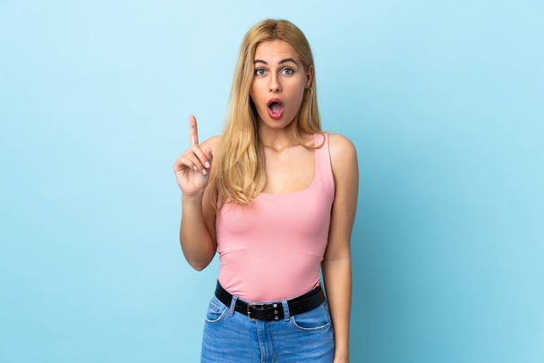 Mladá uruguayská blondýna přes izolované modré pozadí s úmyslem realizovat řešení a zároveň zvednout prst nahoru - Fotografie, Obrázek