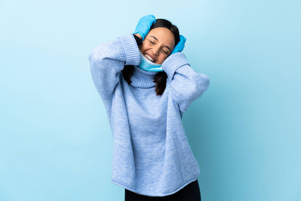 jong brunette gemengd ras vrouw beschermen tegen de coronavirus met een masker en handschoenen over geïsoleerde blauwe achtergrond lachen - Foto, afbeelding