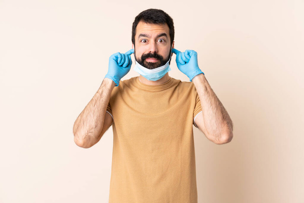 Kaukasischer Mann mit Bart, der sich vor dem Coronavirus schützt, mit Maske und Handschuhen über isoliertem Hintergrund frustriert und mit verdeckenden Ohren - Foto, Bild