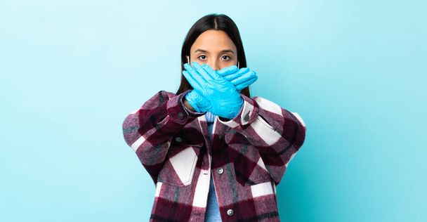 Joven morena de raza mixta protegiendo del coronavirus con una máscara y guantes sobre fondo azul aislado haciendo gesto de stop con la mano para detener un acto
 - Foto, Imagen