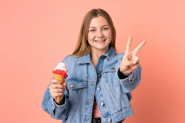 Teenager Ukrainian girl with a cornet ice cream isolated on pink background smiling and showing victory sign - Photo, Image