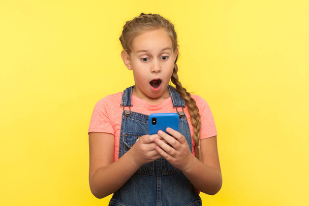 Portrait of shocked little girl reading surprising message on cellphone, chatting on social network, amazed by good mobile application for children. indoor studio shot isolated on yellow background - Zdjęcie, obraz