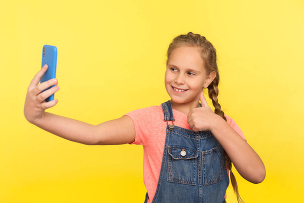 Excellent cellular. Portrait of adorable little girl in denim overalls gesturing thumbs up while communicating with parents on video call, taking selfie on mobile phone. indoor studio shot, isolated - Photo, image