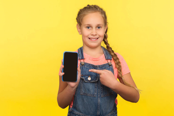Portrait of nice little girl with braid in denim overalls pointing at screen mock up, child showing mobile phone, advertising application and smiling. indoor studio shot isolated on yellow background - 写真・画像