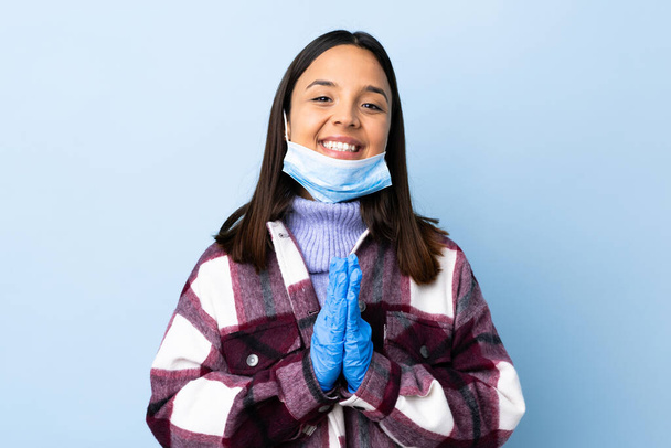 Νεαρή μελαχρινή μικτή φυλετική γυναίκα που προστατεύει από το coronavirus με μάσκα και γάντια πάνω από απομονωμένο μπλε φόντο κρατά την παλάμη μαζί. Κάποιος ζητάει κάτι. - Φωτογραφία, εικόνα