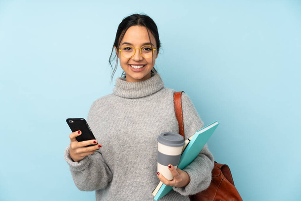 Giovane donna razza mista andando a scuola isolato su sfondo blu con caffè da portare via e un cellulare - Foto, immagini