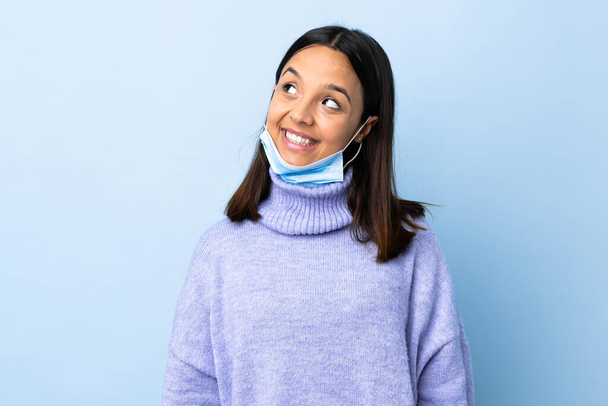 Young brunette mixed race woman protecting from the coronavirus with a mask and gloves over isolated blue background thinking an idea while looking up - Photo, Image