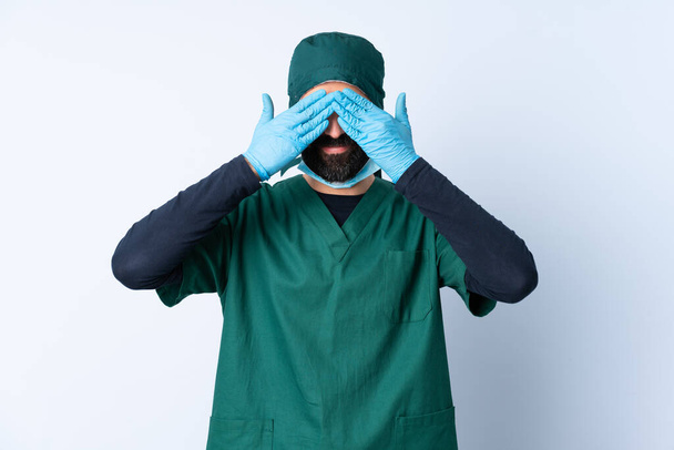 Chirurg man in groen uniform over geïsoleerde achtergrond bedekken ogen door handen en glimlachen - Foto, afbeelding