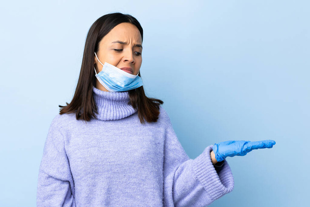 Junge brünette Mischlingshündin schützt sich vor dem Coronavirus mit Maske und Handschuhen über isoliertem blauem Hintergrund und hält Kopierraum mit Zweifeln - Foto, Bild