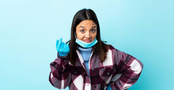 Молодая брюнетка смешанной расы женщина защищает от коронавируса в маске и перчатках на изолированном синем фоне делает итальянский жест
 - Фото, изображение
