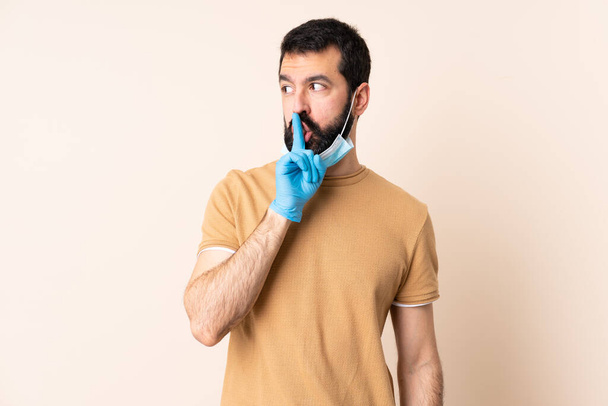 Caucasian man with beard protecting from the coronavirus with a mask and gloves over isolated background showing a sign of silence gesture putting finger in mouth - Fotó, kép