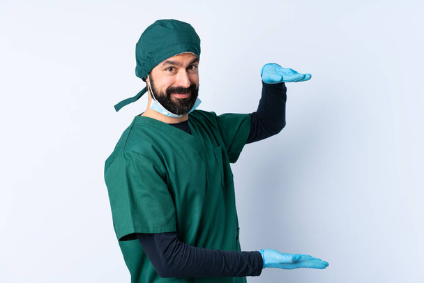 Surgeon man in green uniform over isolated background holding copyspace to insert an ad - Fotografie, Obrázek