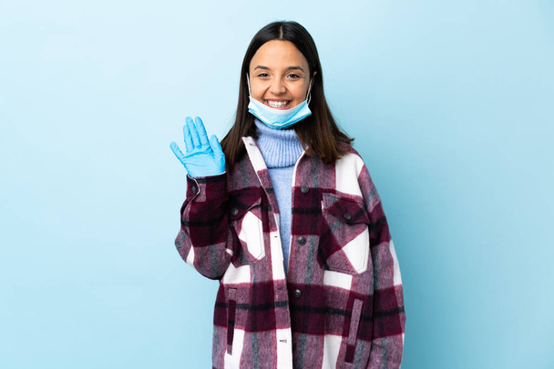 Jovem morena mista mulher protegendo do coronavírus com uma máscara e luvas sobre fundo azul isolado saudando com a mão com expressão feliz
 - Foto, Imagem