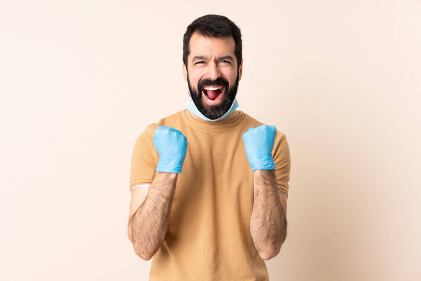 Caucasian man with beard protecting from the coronavirus with a mask and gloves over isolated background frustrated by a bad situation - Фото, изображение