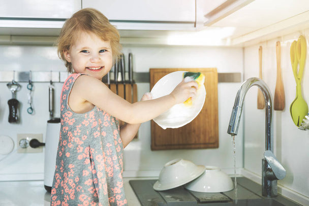 маленькая девочка моет посуду с губкой в кухонной раковине дома
 - Фото, изображение