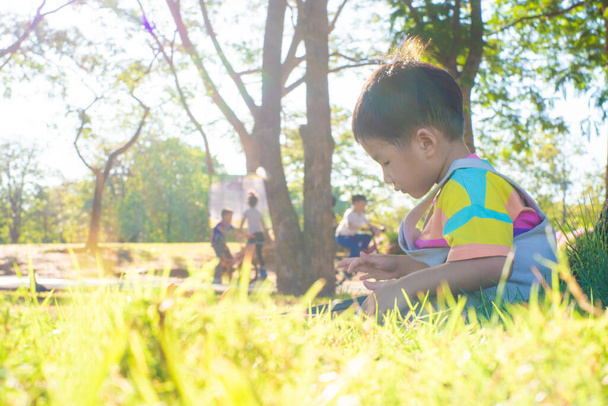 Αξιολάτρευτο ασιατικό αγόρι που κάθεται κάτω από το δέντρο χρήσης tablet τεχνολογία υπολογιστών μαθαίνοντας στο πάρκο ηλιοβασίλεμα φως - Φωτογραφία, εικόνα