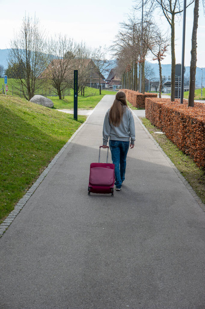 Young Caucasian woman walking on a sidewalk dragging her roller luggage case viewed from behind unrecognizable 2020 - Photo, Image