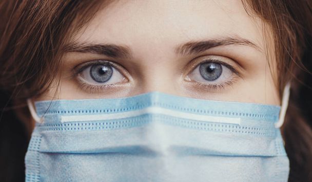 sad eyes of serious young woman face in a protective medical mask on colored studio background, girl looking at the camera, concept of health and quarantine measures - Fotoğraf, Görsel