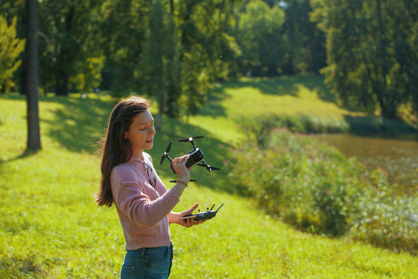 Young woman in a clearing in a park launches a drone, holding an aircraft in one hand and a control panel in the other. - Foto, Imagem