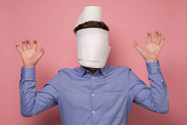 Man in blue shirt wrapped his face in a paper as medical mask trying to protect himself from coronavirus. Studio shot on pink wall. - Photo, Image