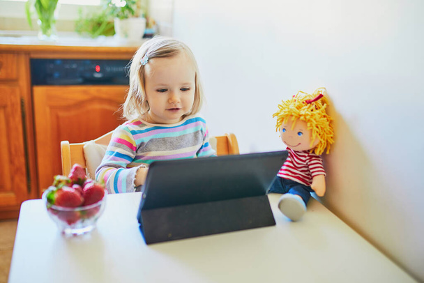 Αξιολάτρευτο κοριτσάκι τρώει φρέσκιες φράουλες και χρησιμοποιεί δισκίο. Παιδί που βλέπει κινούμενα σχέδια ενώ τρώει. Ψηφιακές τεχνολογίες στην εκπαίδευση των παιδιών - Φωτογραφία, εικόνα