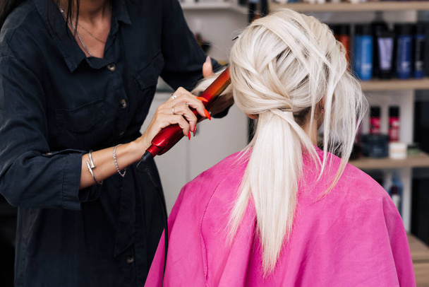 Руки парикмахера с утюгом делают волосы блондинки - Фото, изображение