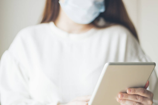 Tıbbi maskeyle korunan genç bir kadın, çalışma ve bilgi için tablet kullanıyor, COVID-19, koronavirüs, seçici odaklanma - Fotoğraf, Görsel
