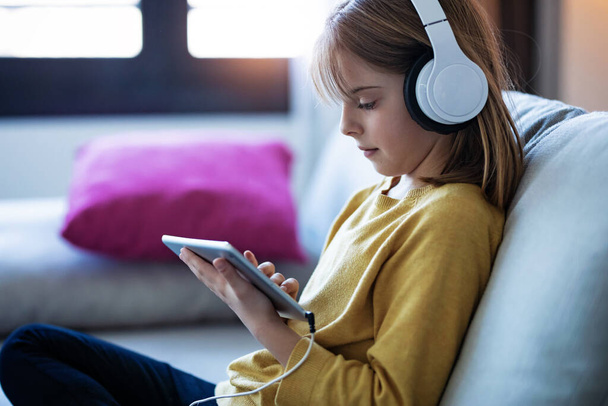 Shot of little girl listening to music with headphones and digital tablet while sitting on sofa at home. - Photo, image