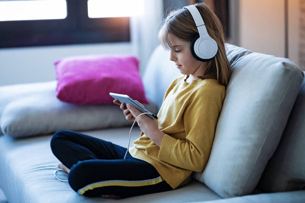 Shot of little girl listening to music with headphones and digital tablet while sitting on sofa at home. - Fotografie, Obrázek